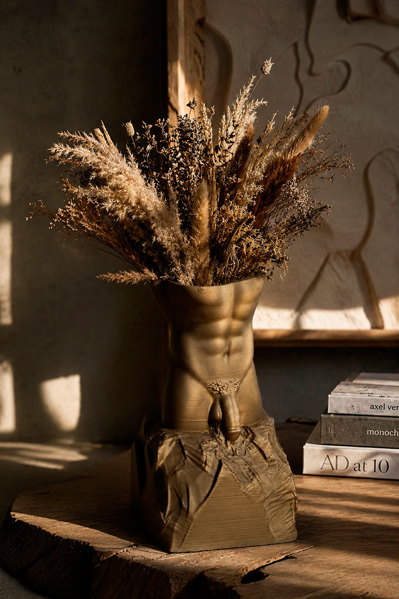 Bronze male torso sculptural vase filled with dried botanical arrangement, displayed on a wooden table in a rustic contemporary interior with warm natural lighting.