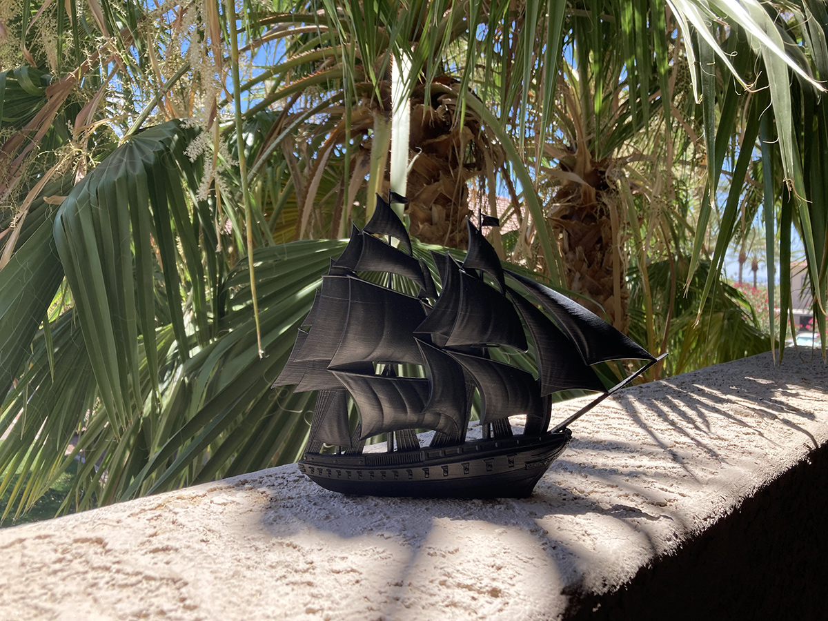Black sailing ship sculpture placed outdoors with palm leaves and natural sunlight, highlighting contrast and silhouette